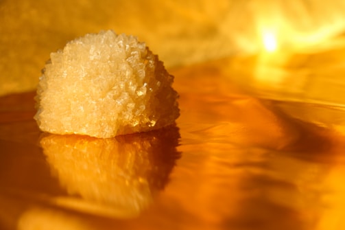 A close-up of a rough crystal or mineral with a golden yellow hue, resting on a reflective golden surface that creates a warm, shimmering effect. The lighting adds a glowing, ambient warmth, highlighting the texture and facets of the crystal.