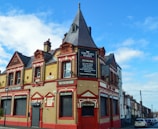 A traditional brick building with a combination of Victorian architectural elements, featuring red and beige tones. The upper part of the structure includes ornate detailing and a clock tower-like peak. Several signs advertise it as a bed and breakfast called 'The King Harry' with amenities such as a function room. The building is located on a street lined with similar row houses.