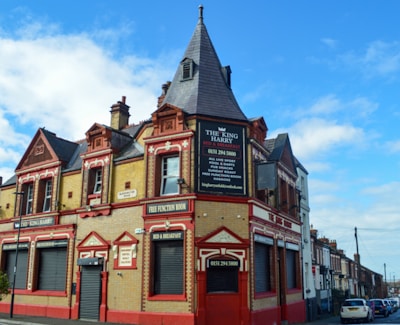 A traditional brick building with a combination of Victorian architectural elements, featuring red and beige tones. The upper part of the structure includes ornate detailing and a clock tower-like peak. Several signs advertise it as a bed and breakfast called 'The King Harry' with amenities such as a function room. The building is located on a street lined with similar row houses.