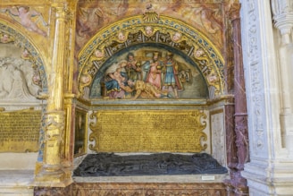 An ornate, historical relief sculpture featuring a religious scene with several figures, likely depicting a biblical event. The artwork is richly decorated with intricate carvings, and the frame includes numerous cherubic faces. There are Latin inscriptions below the scene, alongside an effigy on the tomb.