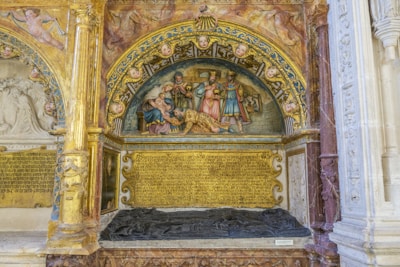 An ornate, historical relief sculpture featuring a religious scene with several figures, likely depicting a biblical event. The artwork is richly decorated with intricate carvings, and the frame includes numerous cherubic faces. There are Latin inscriptions below the scene, alongside an effigy on the tomb.
