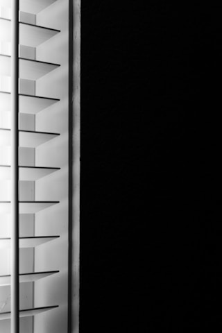 A close-up view of window blinds with horizontal slats, partially open, allowing minimal light to pass through against a dark background.
