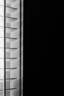 A close-up view of window blinds with horizontal slats, partially open, allowing minimal light to pass through against a dark background.