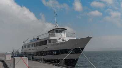 A large ferry named 'NORTHERN SPIRIT I' is docked at a wharf. The vessel is painted white with black accents and features multiple decks with outdoor and indoor areas. The sky is partly cloudy, and the water is calm, with another smaller vessel visible in the distance.