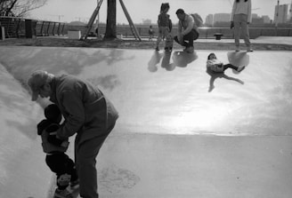 A man assists a young child wearing protective gear on a skate ramp. Another child is lying down on the ramp nearby, while two other adults and a child observe from the top of the ramp. The setting appears to be an outdoor park area with trees and buildings in the background.