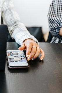 A hand with manicured nails and rings is interacting with a smartphone placed on a dark table. The smartphone screen displays a grid of images, suggesting the use of a social media or photo gallery app. The person is wearing a plaid long-sleeve shirt, and another person is partially visible beside them, also wearing a checkered shirt.