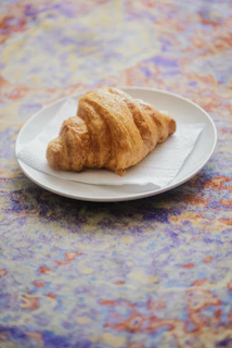 Golden croissant resting on a soft beige linen cloth with warm morning light.