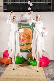 Two performers on stilts wearing elaborate white costumes and golden masks are standing in front of a large can labeled 'Social Cut' with colorful bean bags around them. They appear to be entertaining with bubbles in the air.