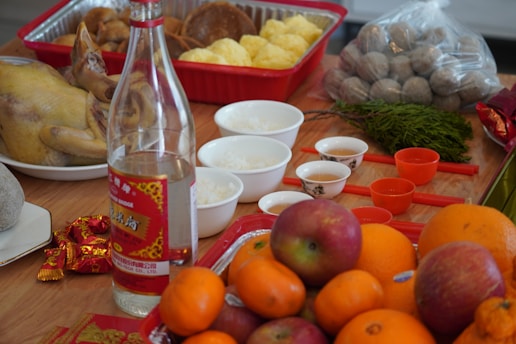 An assortment of food items arranged on a wooden surface, including a whole cooked chicken, a variety of fruits such as oranges and apples, a bottle of alcohol, several small bowls with rice and three small cups, likely for tea. There are also some packaged snacks and eggs in the background.