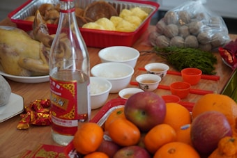 An assortment of food items arranged on a wooden surface, including a whole cooked chicken, a variety of fruits such as oranges and apples, a bottle of alcohol, several small bowls with rice and three small cups, likely for tea. There are also some packaged snacks and eggs in the background.