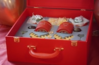 A luxurious red gift box is open, revealing a neatly arranged set of objects including two red containers with golden designs, possibly featuring traditional Chinese motifs. There are also other items, such as wrapped goods and decorative wooden pieces, all placed in custom compartments. The box itself has a red handle and gold clasps, adding to the elegant presentation.