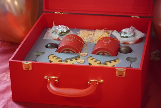 A luxurious red gift box is open, revealing a neatly arranged set of objects including two red containers with golden designs, possibly featuring traditional Chinese motifs. There are also other items, such as wrapped goods and decorative wooden pieces, all placed in custom compartments. The box itself has a red handle and gold clasps, adding to the elegant presentation.