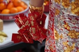 A close-up of an intricately embroidered garment with red and gold patterns. The person is holding a cluster of bright red envelopes featuring gold Chinese characters. There is a background of oranges on a red tray, which hints at a festive setting.