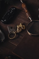 A wooden surface holds a collection of items including a folding knife with a dark handle and metallic elements, a set of keys attached to a gold-colored dragon keychain, a magnifying glass with a wooden handle, and a closed notebook with a textured cover.