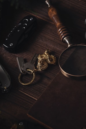 A wooden surface holds a collection of items including a folding knife with a dark handle and metallic elements, a set of keys attached to a gold-colored dragon keychain, a magnifying glass with a wooden handle, and a closed notebook with a textured cover.