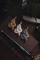 Three serpentine dragon-shaped pendants in gold, silver, and dark metal are placed on an open leather notebook. Next to the notebook, there is a black tactical pen, a silver folding knife, and a black folding knife.