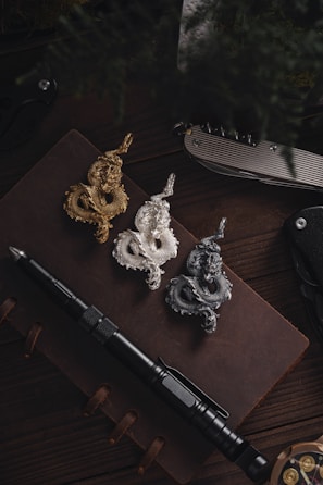 Three serpentine dragon-shaped pendants in gold, silver, and dark metal are placed on an open leather notebook. Next to the notebook, there is a black tactical pen, a silver folding knife, and a black folding knife.