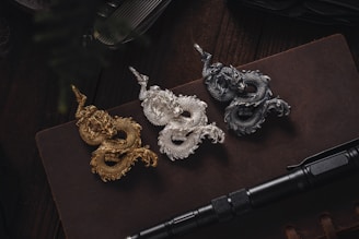 Three intricately designed dragon figurines in gold, silver, and bronze are arranged on a dark book or notebook. Below the figurines, a black tactical pen is placed horizontally. The setting is dimly lit, adding a sense of mystery and elegance.