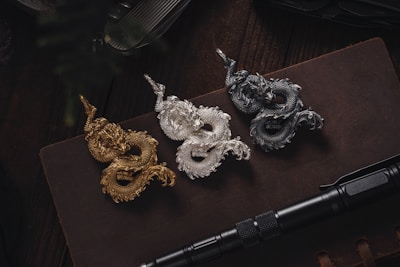Three intricately designed dragon figurines in gold, silver, and bronze are arranged on a dark book or notebook. Below the figurines, a black tactical pen is placed horizontally. The setting is dimly lit, adding a sense of mystery and elegance.