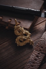 A golden dragon figurine is placed on a dark wooden surface next to a leather-bound notebook and a black flashlight. The scene is illuminated with soft lighting, highlighting the intricate details of the dragon and the texture of the leather.