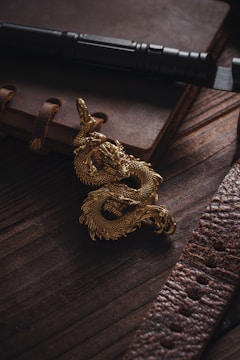 A golden dragon figurine is placed on a dark wooden surface next to a leather-bound notebook and a black flashlight. The scene is illuminated with soft lighting, highlighting the intricate details of the dragon and the texture of the leather.