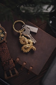 A close-up of a brown leather-bound notebook with a rugged texture. On top of the notebook, there is a set of keys attached to a golden dragon keychain. The background includes a watch with a leather strap and metallic bezel, as well as a folding knife with a textured handle.