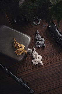 Three intricately designed dragon sculptures in gold, silver, and gray are placed on a dark wooden surface. A metal flask is positioned beside them, along with a folding knife and a flashlight. The setting is dimly lit, with a few shadows and a muted tone.
