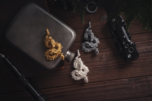 Three intricate dragon-shaped pendants in gold, silver, and bronze are arranged on a wooden surface. Nearby items include a metallic flask, a folding knife, and a pen, creating a rustic and rugged atmosphere.