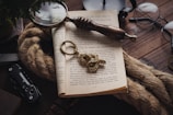 A wooden table holds an array of objects, including an open book with text, a magnifying glass with a wooden handle resting on the pages, and a thick rope beside the book. Nearby, a pair of eyeglasses and an ornate, metal keychain are visible. The setting suggests a vintage or explorer theme.