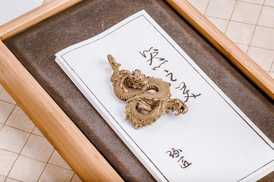 A gold-colored dragon ornament rests on a piece of white paper with Asian characters written on it. The setup is elegantly placed in a wooden box with a textured gray interior. The paper is neatly framed, highlighting the intricate details of the dragon's design.