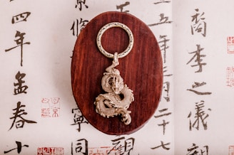A detailed and intricately designed dragon keychain rests on an oval wooden stand. The background features traditional East Asian calligraphy, adding an artistic and cultural touch to the composition.