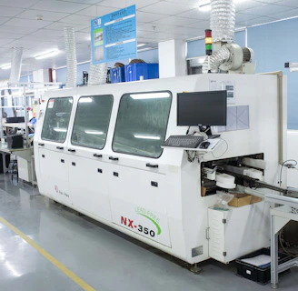 A modern electronics manufacturing facility featuring a large machine with a computer monitor attached. The machine is labeled NX-350 and appears to be part of a production line. There are tables and chairs lined up, with various equipment and components scattered across the surfaces. The setting is well-lit, and the environment is tidy and organized, with a visible air filtering system installed above.