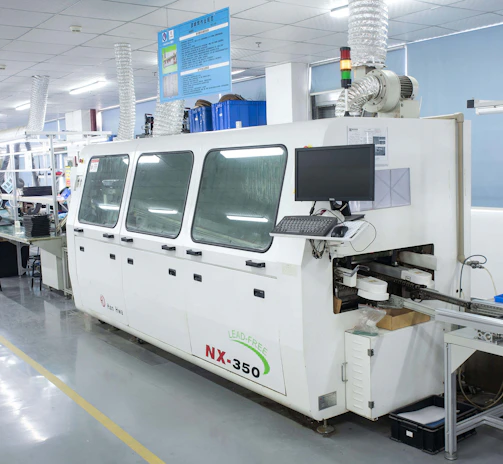 A modern electronics manufacturing facility featuring a large machine with a computer monitor attached. The machine is labeled NX-350 and appears to be part of a production line. There are tables and chairs lined up, with various equipment and components scattered across the surfaces. The setting is well-lit, and the environment is tidy and organized, with a visible air filtering system installed above.