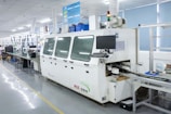 A modern electronics manufacturing facility featuring a large machine with a computer monitor attached. The machine is labeled NX-350 and appears to be part of a production line. There are tables and chairs lined up, with various equipment and components scattered across the surfaces. The setting is well-lit, and the environment is tidy and organized, with a visible air filtering system installed above.