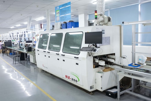 A modern electronics manufacturing facility featuring a large machine with a computer monitor attached. The machine is labeled NX-350 and appears to be part of a production line. There are tables and chairs lined up, with various equipment and components scattered across the surfaces. The setting is well-lit, and the environment is tidy and organized, with a visible air filtering system installed above.