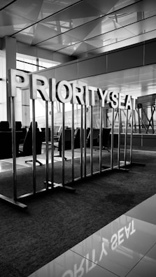 A section of an airport seating area designated as 'Priority Seat.' The section is marked by large metal letters on poles that spell out 'PRIORITY SEAT.' The surrounding area features modern, sleek architecture with large windows and polished surfaces. Chairs can be seen in the background, facing the windows.