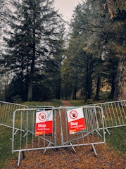 A forested area with a path partially blocked by metal barriers, displaying red warning signs indicating danger. The trees are dense and the atmosphere is somewhat gloomy.