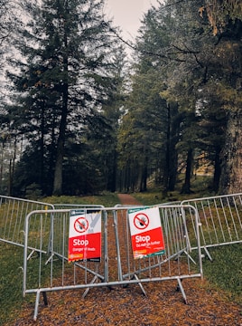 A forested area with a path partially blocked by metal barriers, displaying red warning signs indicating danger. The trees are dense and the atmosphere is somewhat gloomy.