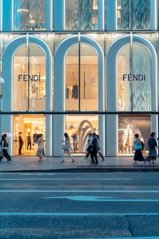 A modern storefront for the brand Fendi features large glass windows with illuminated displays. People walk along the sidewalk in front of the store, showcasing a bustling urban environment. The store's design includes tall, arched sections that are visually striking.