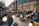 A cozy outdoor space decorated with warm string lights, creating a festive and inviting atmosphere. The area is surrounded by a series of arcaded brick buildings, with people enjoying their time at tables beneath large umbrellas. Plant pots add a touch of greenery to the scene.