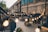 A cozy outdoor space decorated with warm string lights, creating a festive and inviting atmosphere. The area is surrounded by a series of arcaded brick buildings, with people enjoying their time at tables beneath large umbrellas. Plant pots add a touch of greenery to the scene.