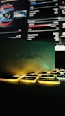 A box labeled 'AirPods Pro' rests partially on a keyboard with illuminated keys. In the background, a computer screen displays detailed graphs and statistics.