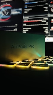 A box labeled 'AirPods Pro' rests partially on a keyboard with illuminated keys. In the background, a computer screen displays detailed graphs and statistics.