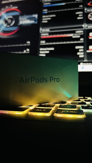 A box labeled 'AirPods Pro' rests partially on a keyboard with illuminated keys. In the background, a computer screen displays detailed graphs and statistics.
