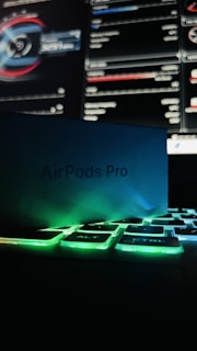 A box labeled 'AirPods Pro' is set against an illuminated keyboard with green backlighting. In the background, a computer screen displays various graphs and data in a dark setting.