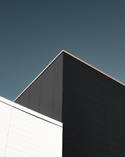 A minimalist architectural structure composed of geometric shapes, featuring right angles and lines. The building has two distinct sections, one in dark grey and the other in white, set against a clear sky.