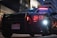 A police car is parked in an urban environment, illuminated by both natural and artificial lights. The vehicle's headlights and red emergency lights are on, and the license plate indicates it is from British Columbia. The cityscape backdrop includes modern buildings with geometric designs.