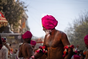 An older man with a white beard and white mustache, wearing a bright pink turban, is prominently featured. He is bare-chested and adorned with colorful cloth bracelets. Other individuals in the background are also wearing pink turbans. Trees and buildings can be seen in the distant background.