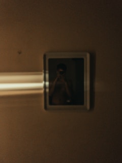 A dimly lit photograph with a reflective surface showing a blurry figure inside a frame. Light streaks cross the left side of the frame, enhancing the mysterious ambiance.