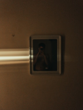 A dimly lit photograph with a reflective surface showing a blurry figure inside a frame. Light streaks cross the left side of the frame, enhancing the mysterious ambiance.